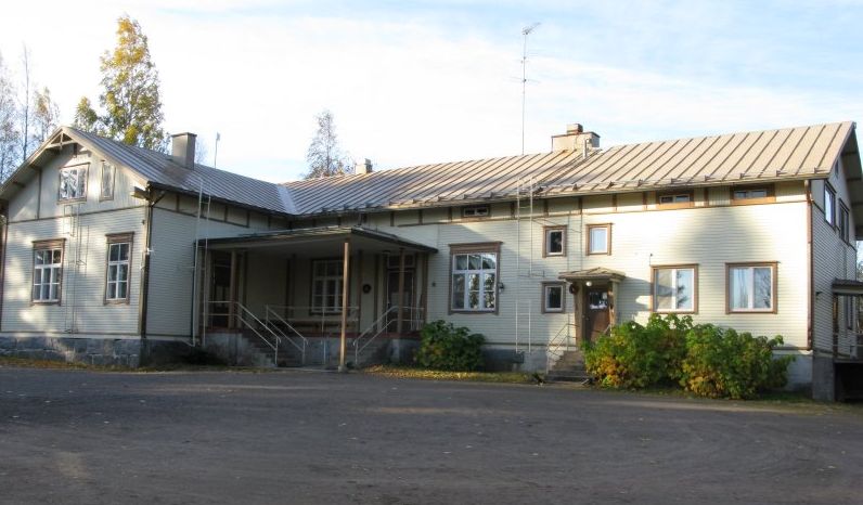 Pitkä yksikerroksinen puurakennus peltikatolla ja useilla ikkunoilla, taustalla pilvinen taivas